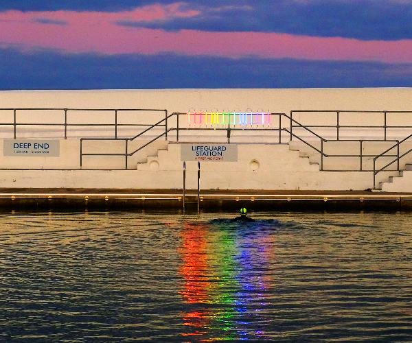 A person swimming through reflections on the water from a colourful neon rainbow glowing on the pool side of the Penzance lido
