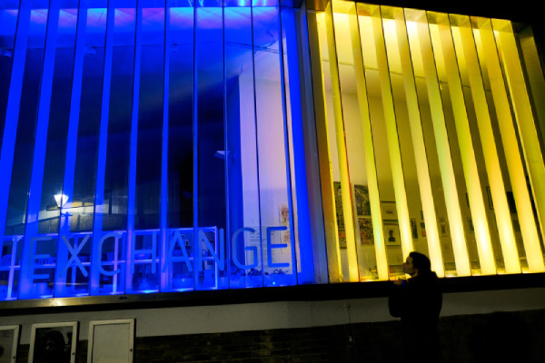 The illuminated facade of the Exchange Gallery in the colours of blue and yellow with a woman looking in